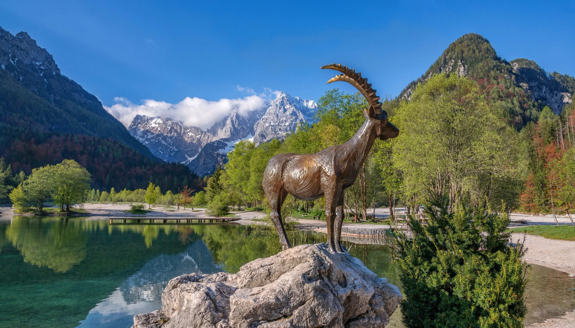 Bronze Zlatorog statue on rock by alpine lake with snow-capped mountains in background.