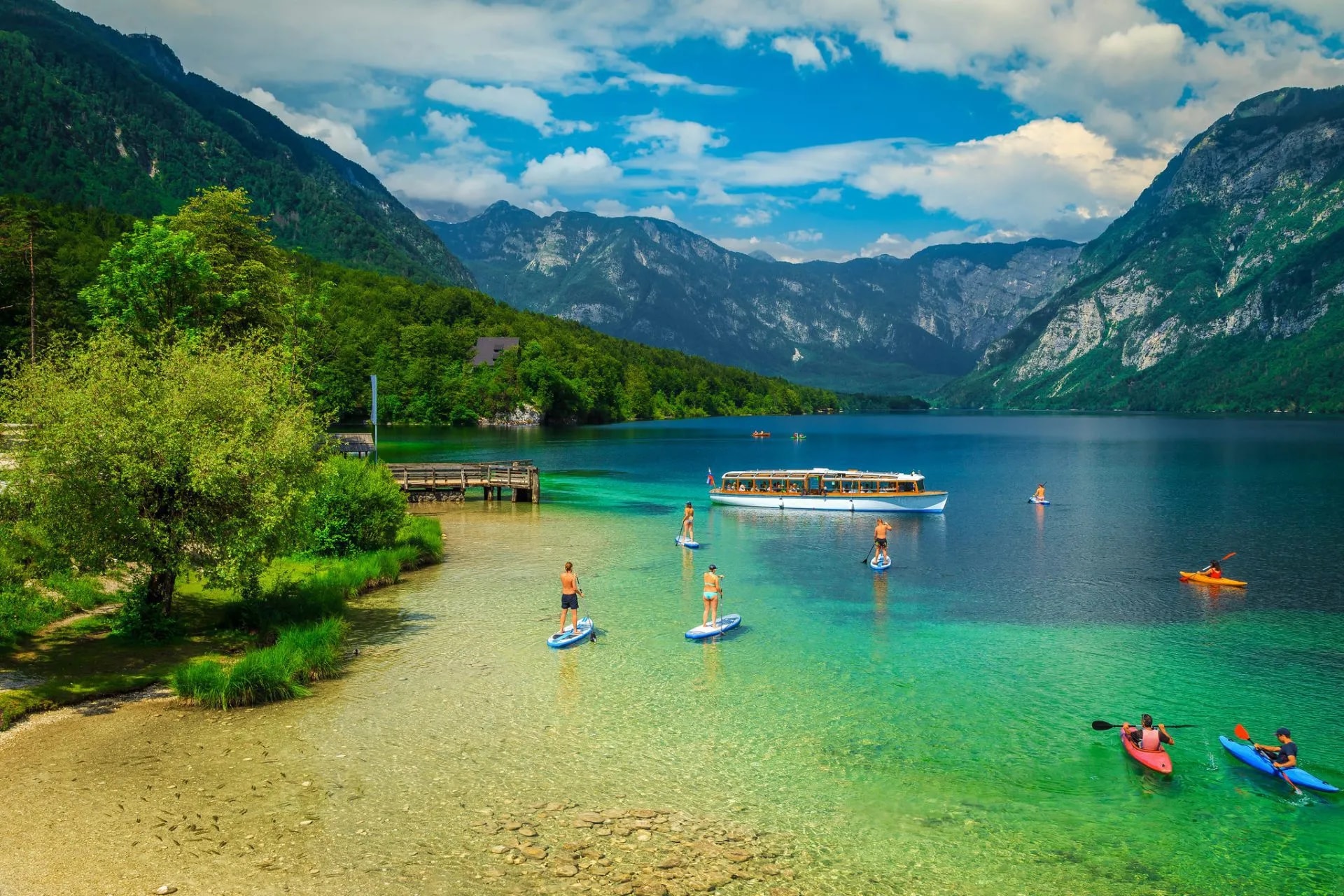 Paddleboarding and kayaking on clear water of Lake Bohinj with a tour boat and mountains.