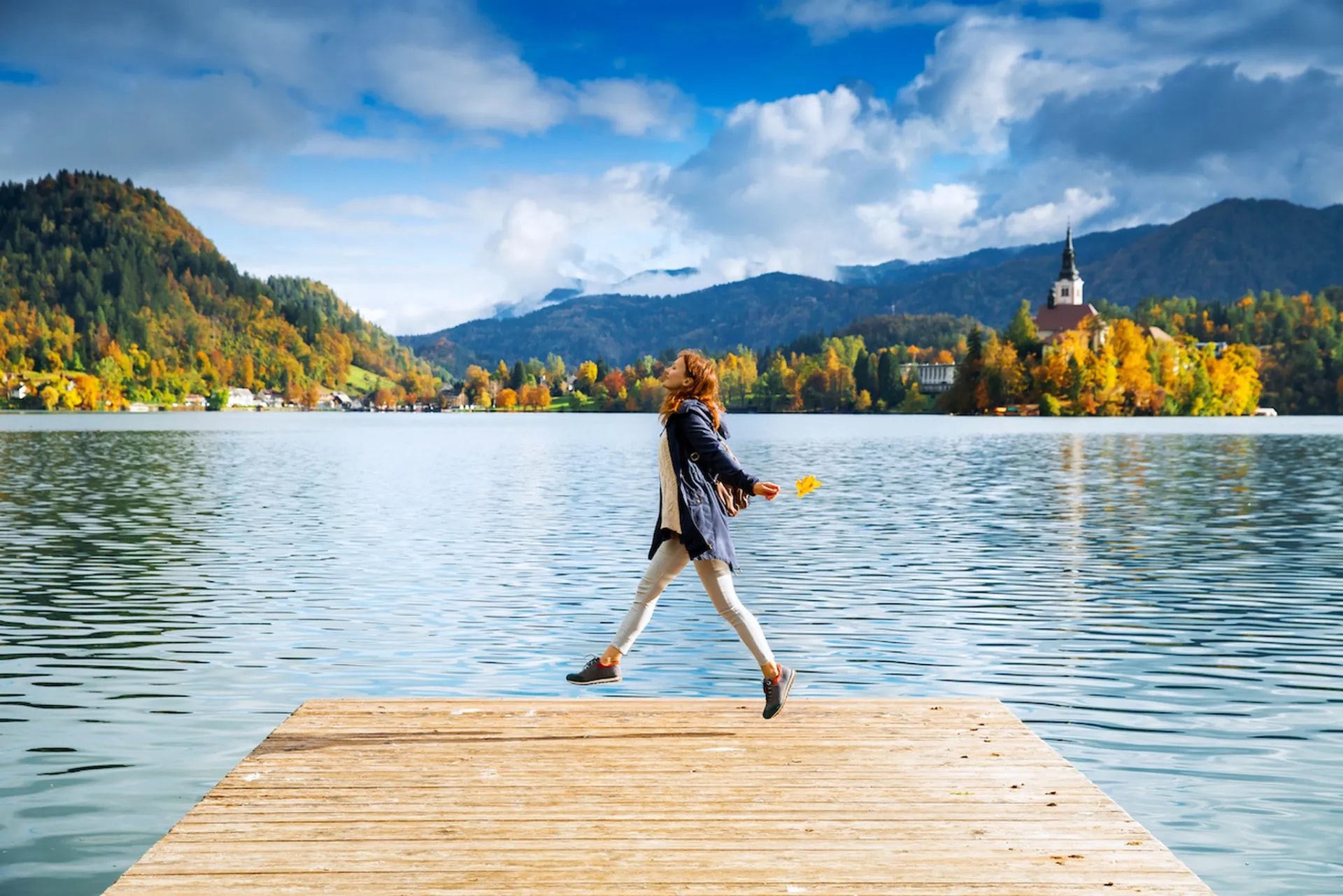 Woman jumping on wooden dock at Lake Bled with island church and autumn mountains