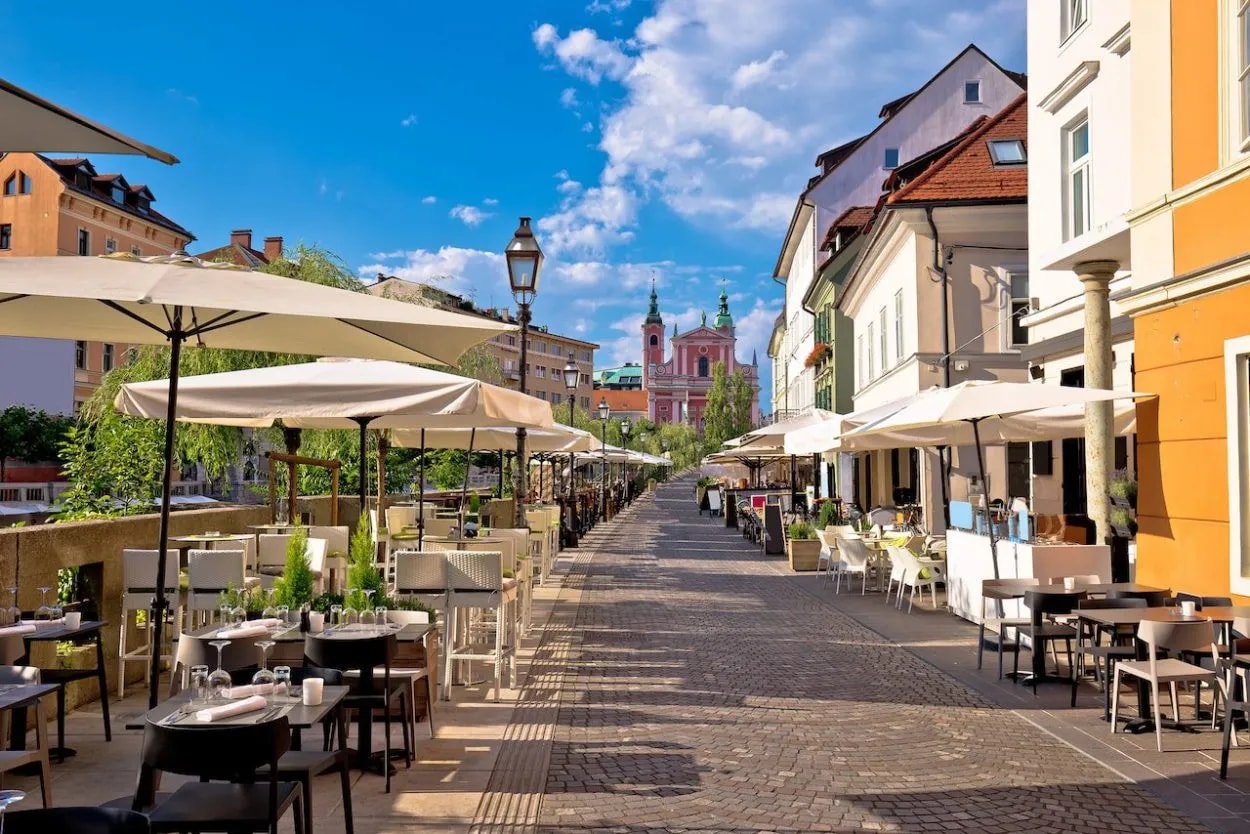 Outdoor cafe seating lines a cobblestone street toward the pink Franciscan Church in Ljubljana.