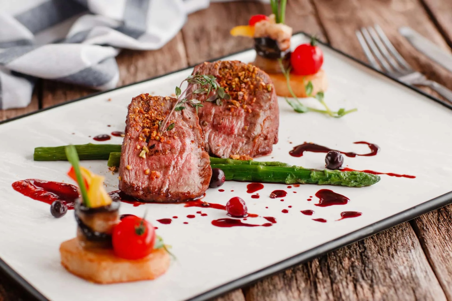 Two medium-rare steak medallions with crust served with asparagus and berry sauce on a white plate.