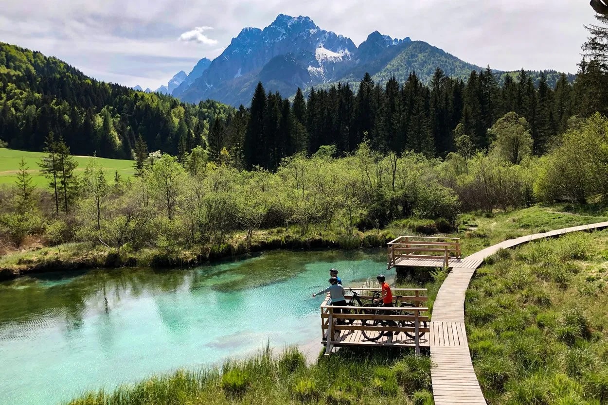 Mountain bikers resting by turquoise spring water with wooden boardwalk and snow-capped peaks.