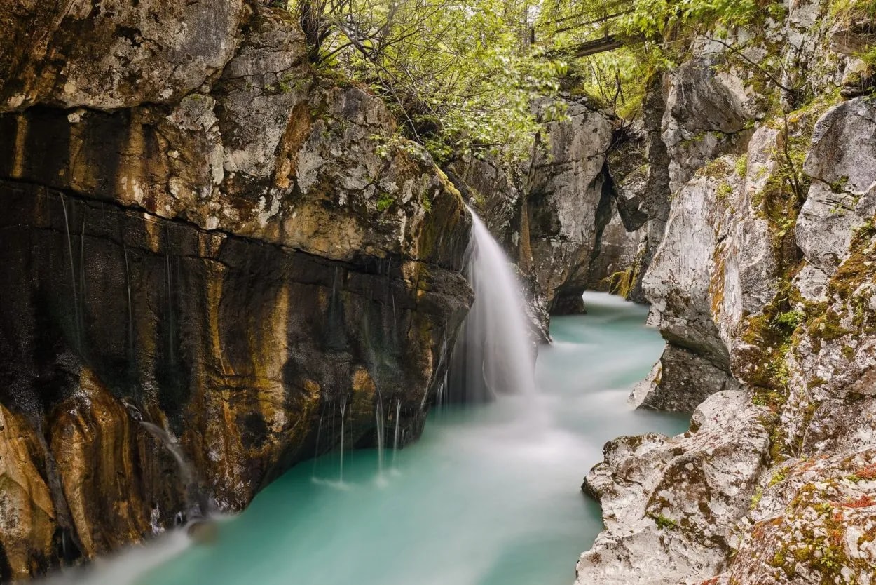 Waterfall cascading into turquoise river in a rocky gorge with lush green foliage.