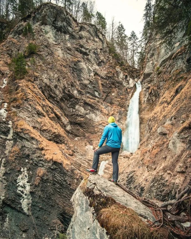Hiker viewing Martuljek waterfall cascading down steep, rocky slopes in a gorge setting.