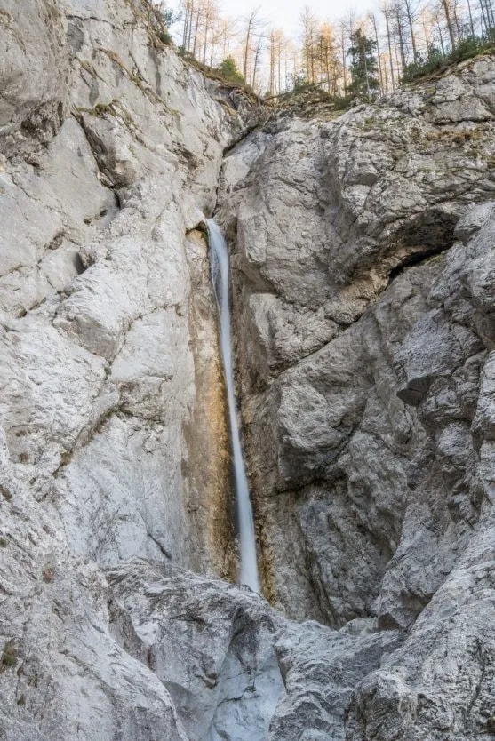 Tall, narrow waterfall cascading down sheer gray rock cliffs with sparse trees at the top.