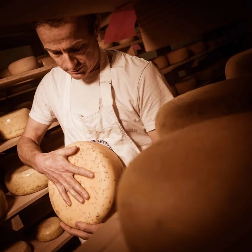 Artisan inspecting a wheel of speckled cheese in a dimly lit cheese aging room with shelves of rounds.