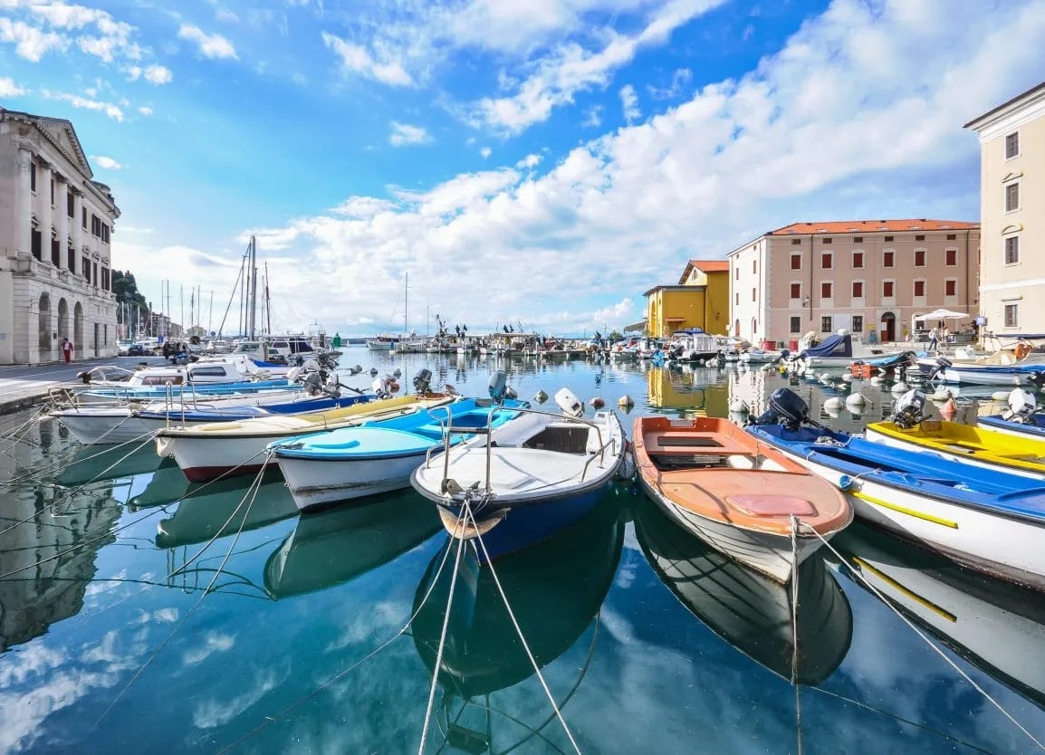 Colorful boats moored in the harbor of Piran under a bright blue, cloudy sky.