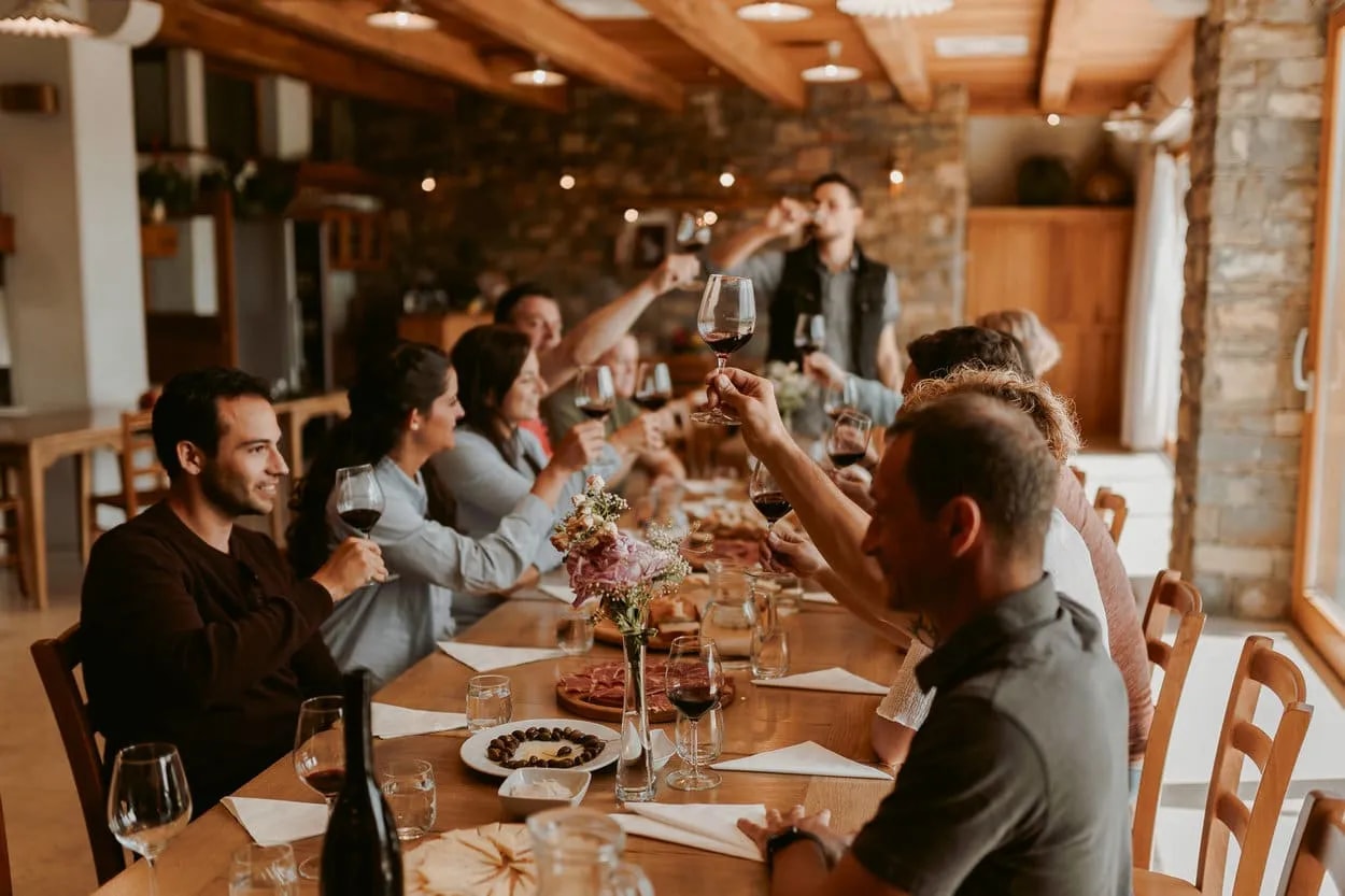 Group toasting with red wine glasses during a meal in a rustic winery or tasting room.