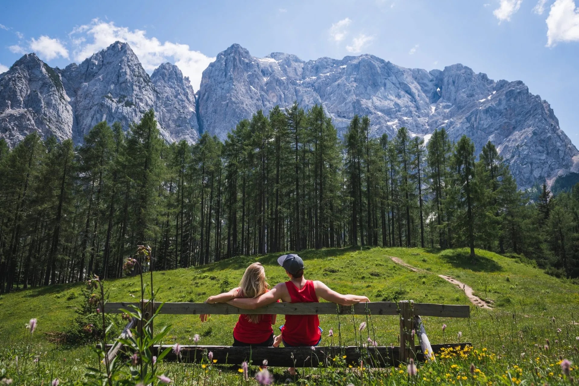 Couple resting on bench viewing Vrsic Pass mountains above pine forest and green meadow.