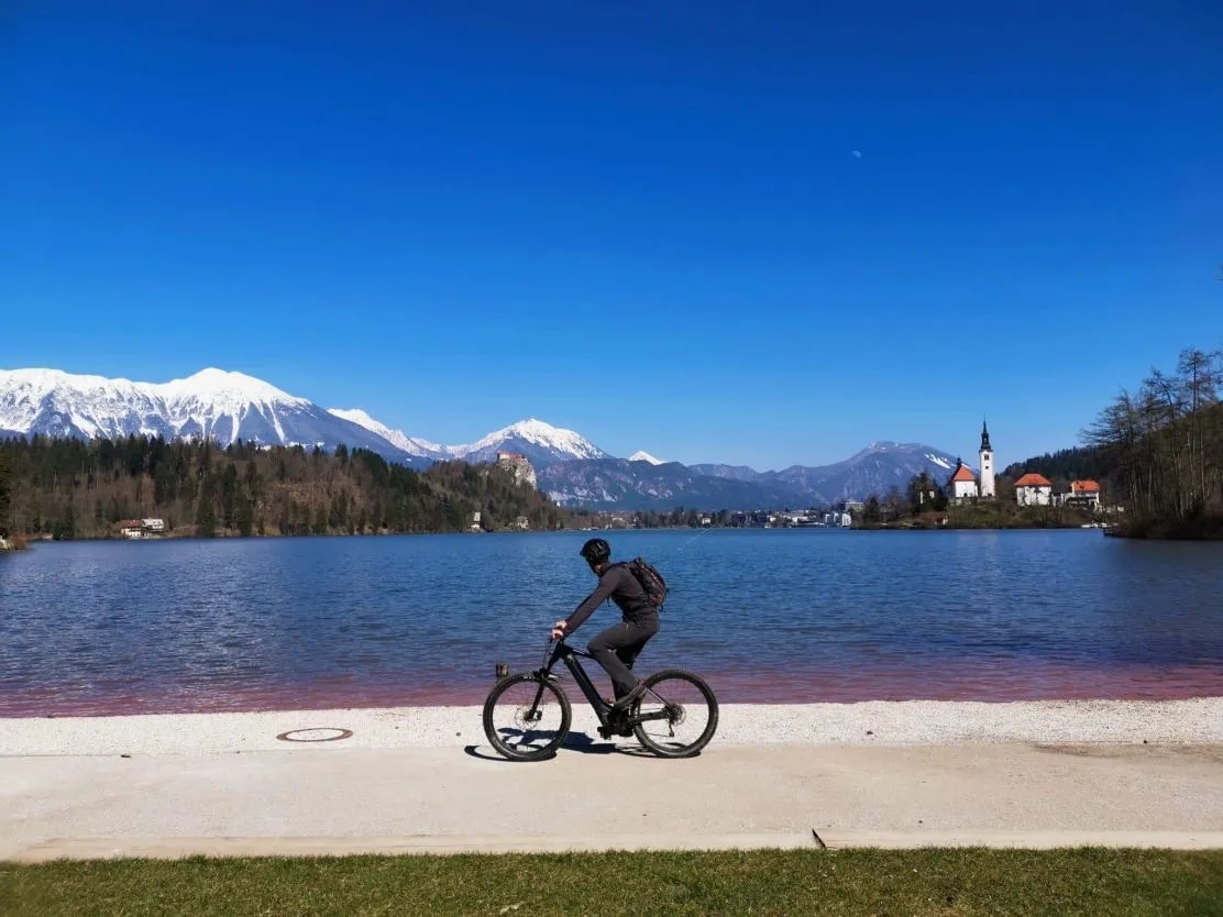 Cyclist riding electric bike by Lake Bled with snow-capped mountains and church island
