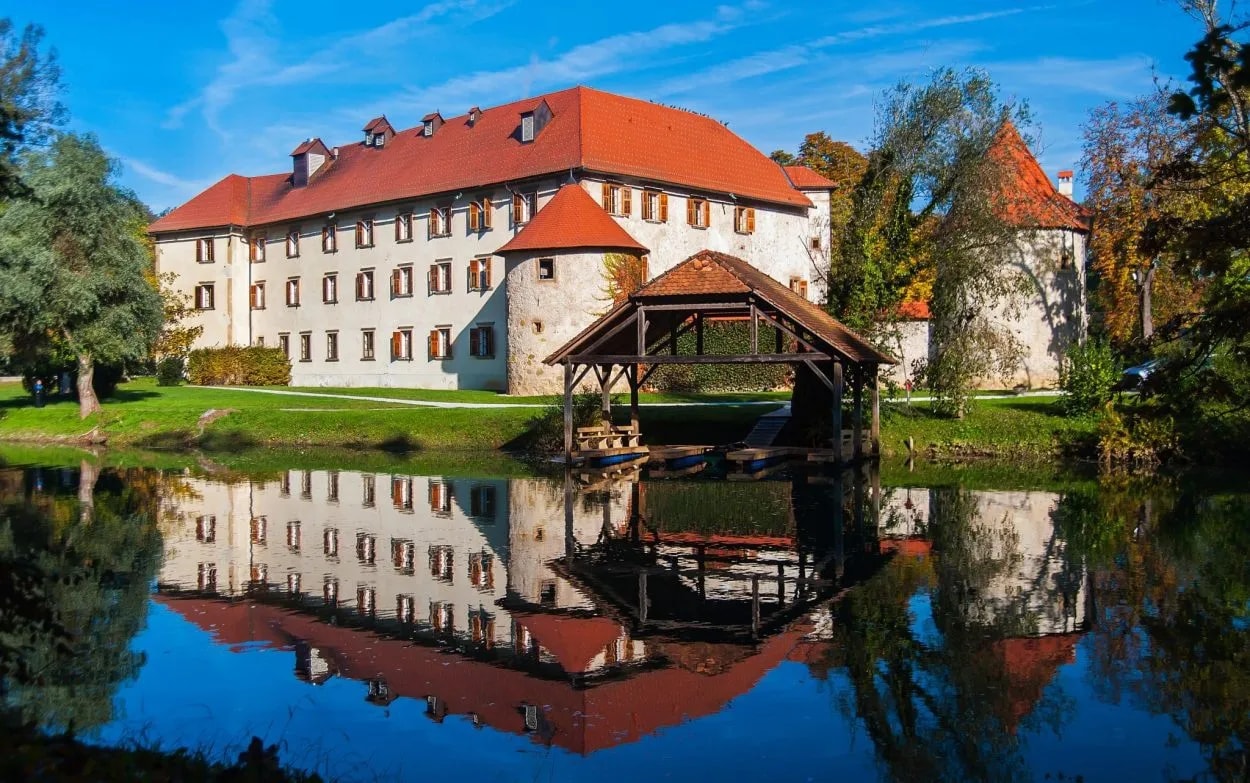 Otočec Castle with red roof reflected in calm water next to a wooden boat dock.