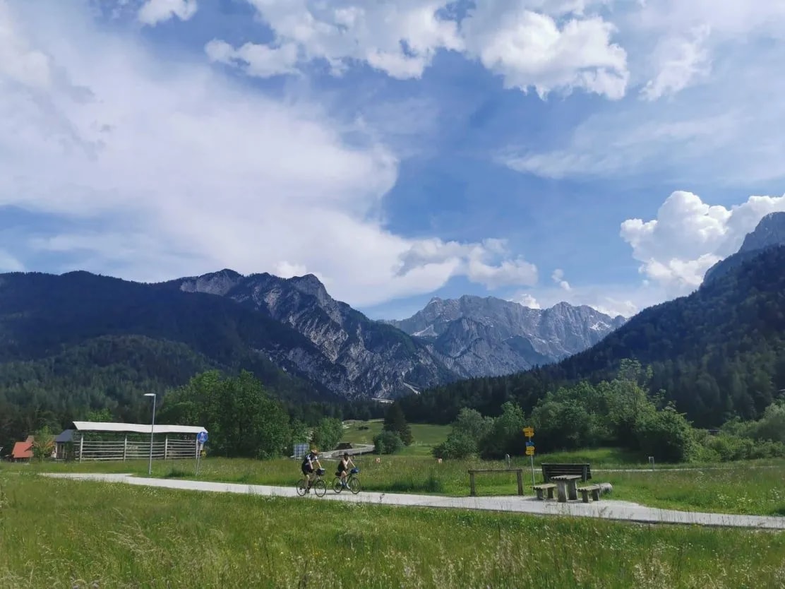 Two people cycling on a path past grassy fields with large mountains under a blue sky.