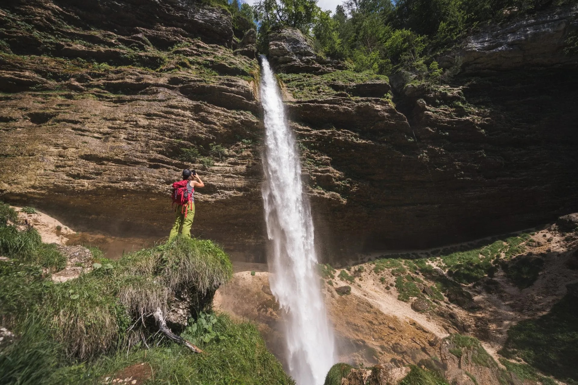 Hiker photographing tall Pericnik Waterfall cascading down a steep, mossy rock face in summer.
