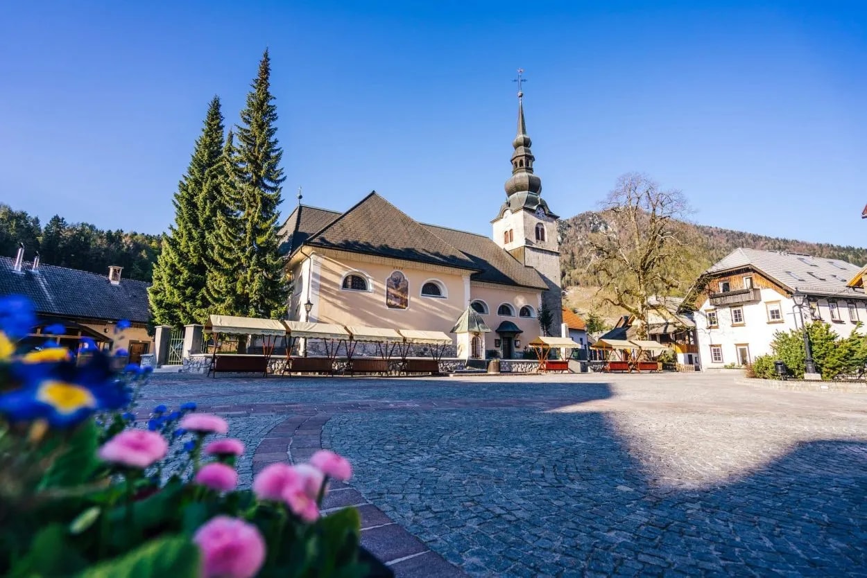 Church with tall spire in Kranjska Gora town square with cobblestones and spring flowers.