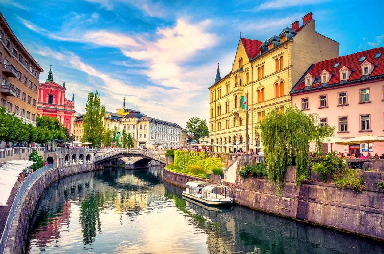 Ljubljana city center river scene with a tour boat, bridge, and colorful historic buildings.