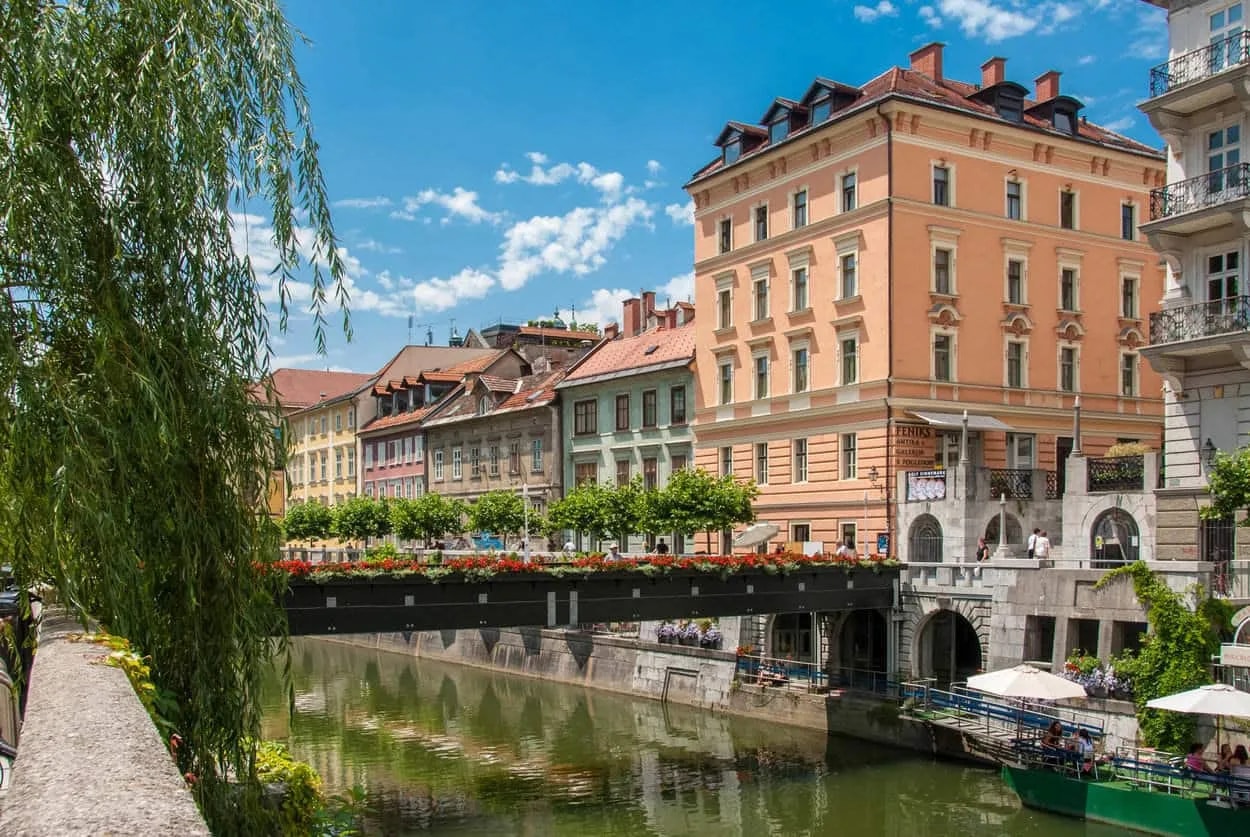 River Ljubljana with historic buildings, a bridge decorated with flowers, and weeping willow in foreground.