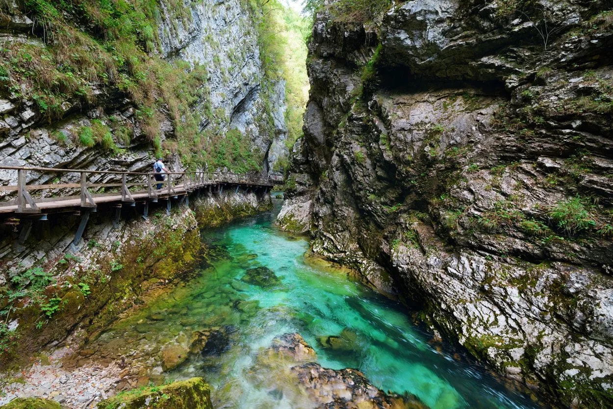 Hiker on wooden walkway above turquoise river in Vintgar Gorge canyon with steep rock walls.