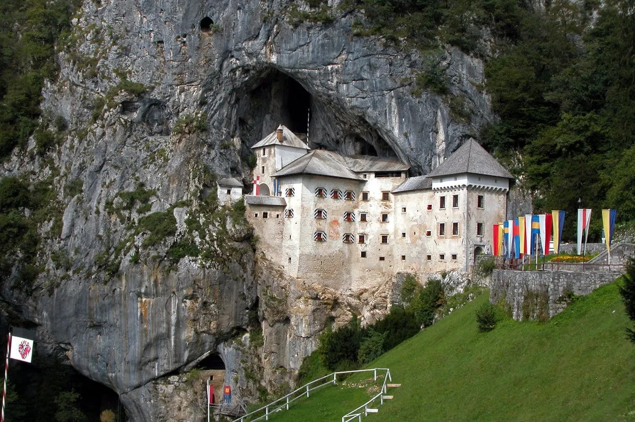Predjama Castle built into a sheer cliff face with a green grassy slope and colorful flags.