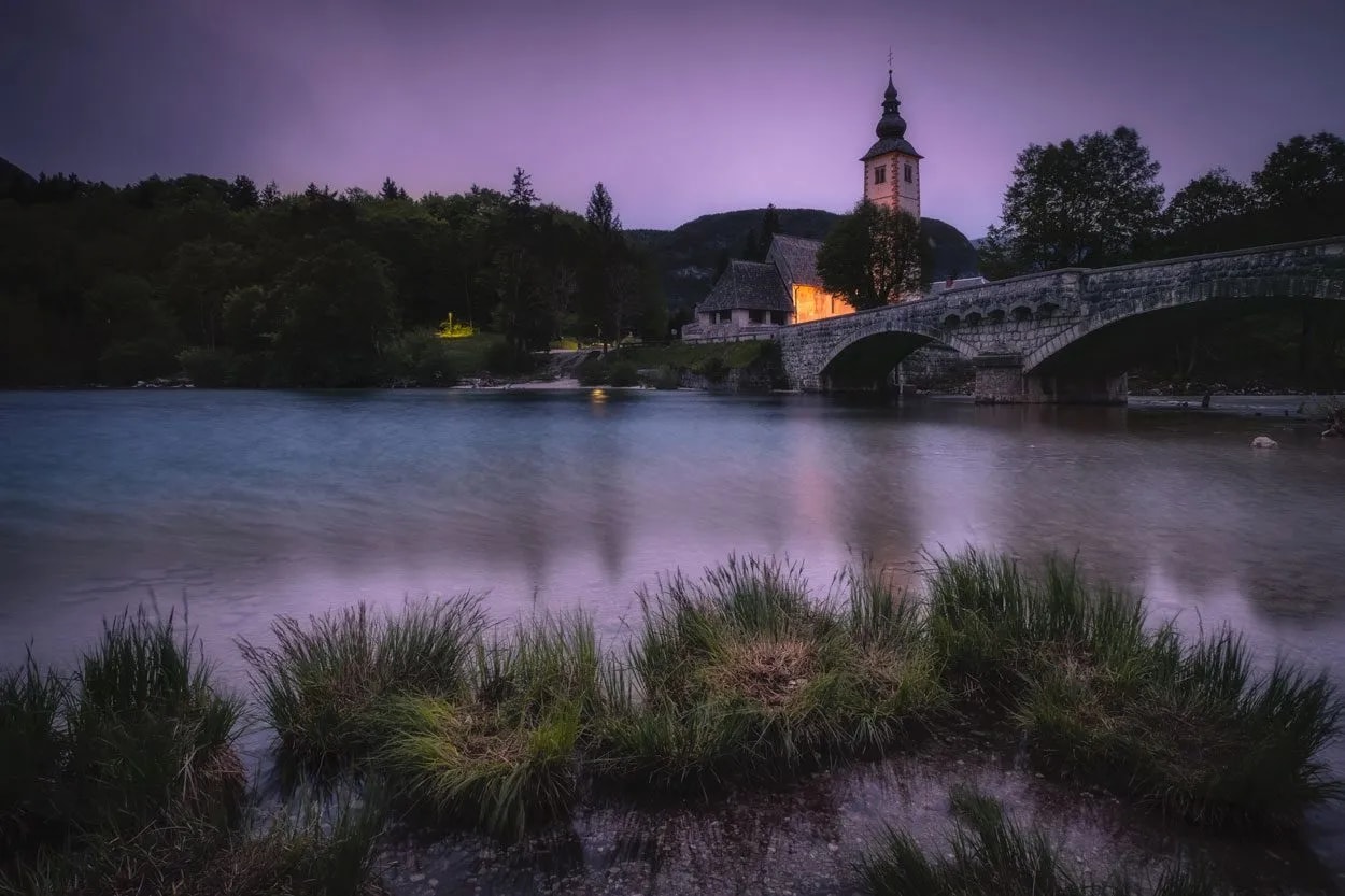 Stone bridge and church tower over river at dusk near Bohinj with foreground grass.