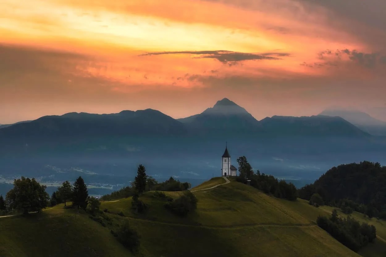 Church on a grassy hill overlooking mountains at sunset in Jamnik, Slovenia