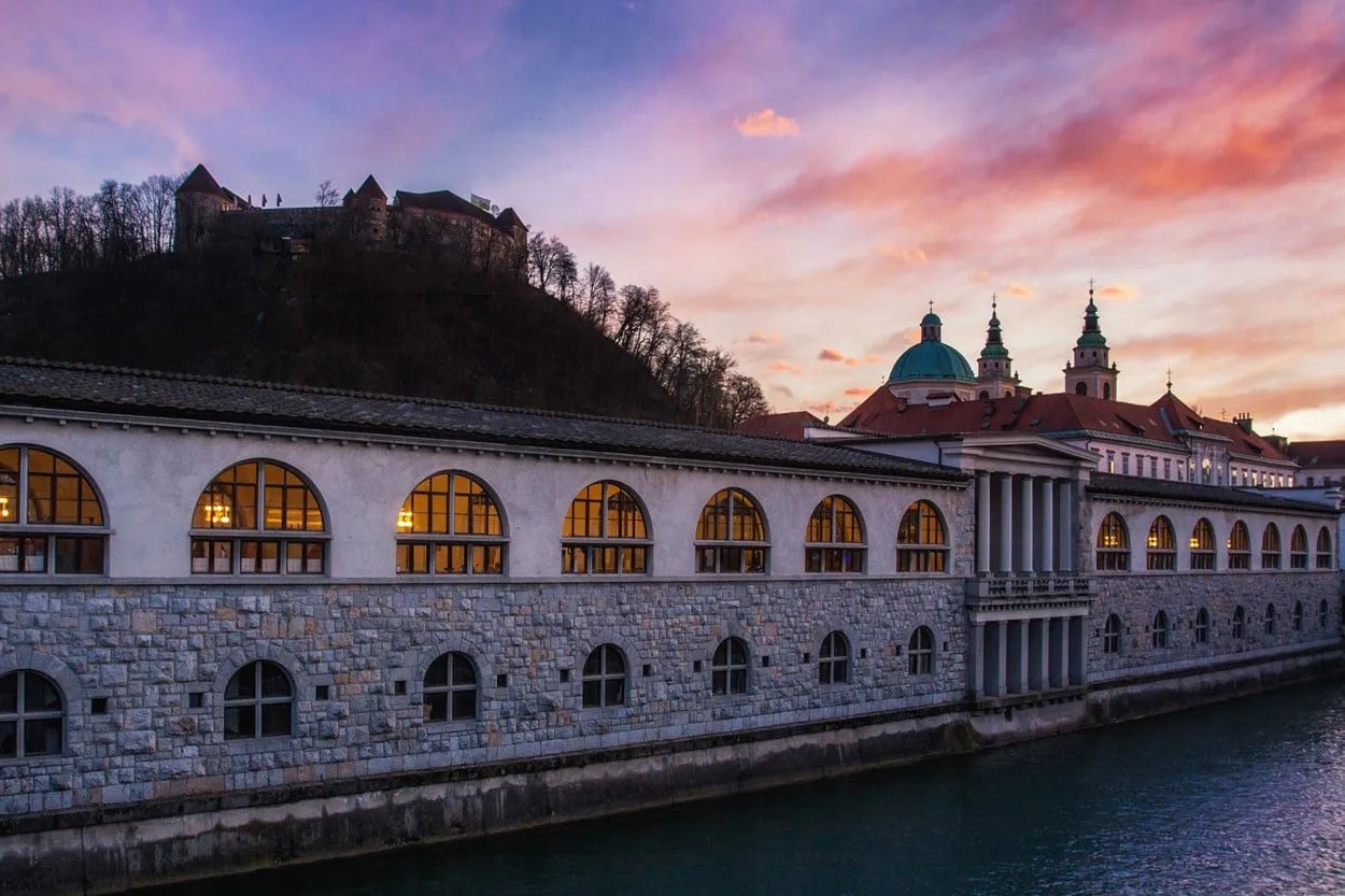 Ljubljana market building by river with castle on hill at sunset