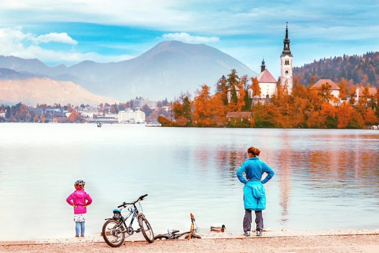 Woman and child with bicycles look at Lake Bled island church with autumn foliage.