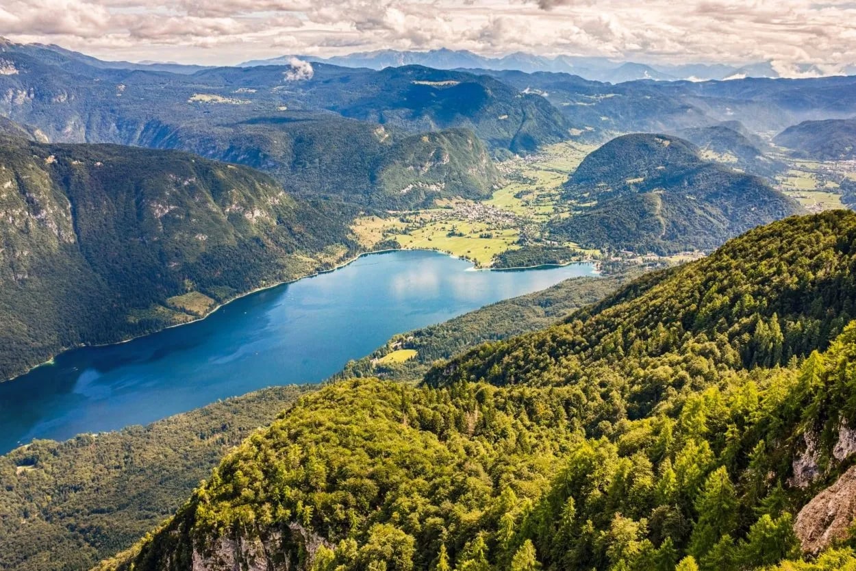 Aerial view of Lake Bohinj surrounded by green mountains and forested slopes under a cloudy sky.