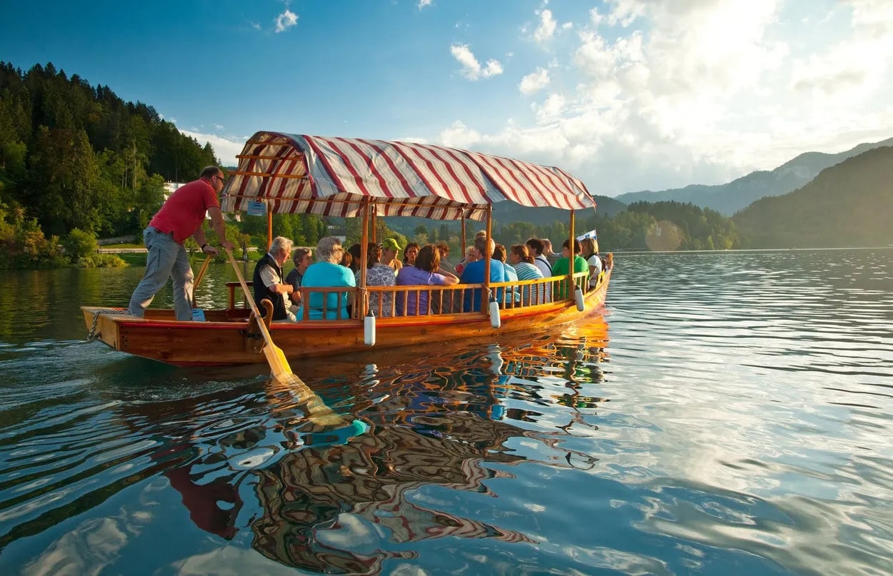 Wooden tourist boat with striped canopy being rowed across Lake Bled with mountains in background