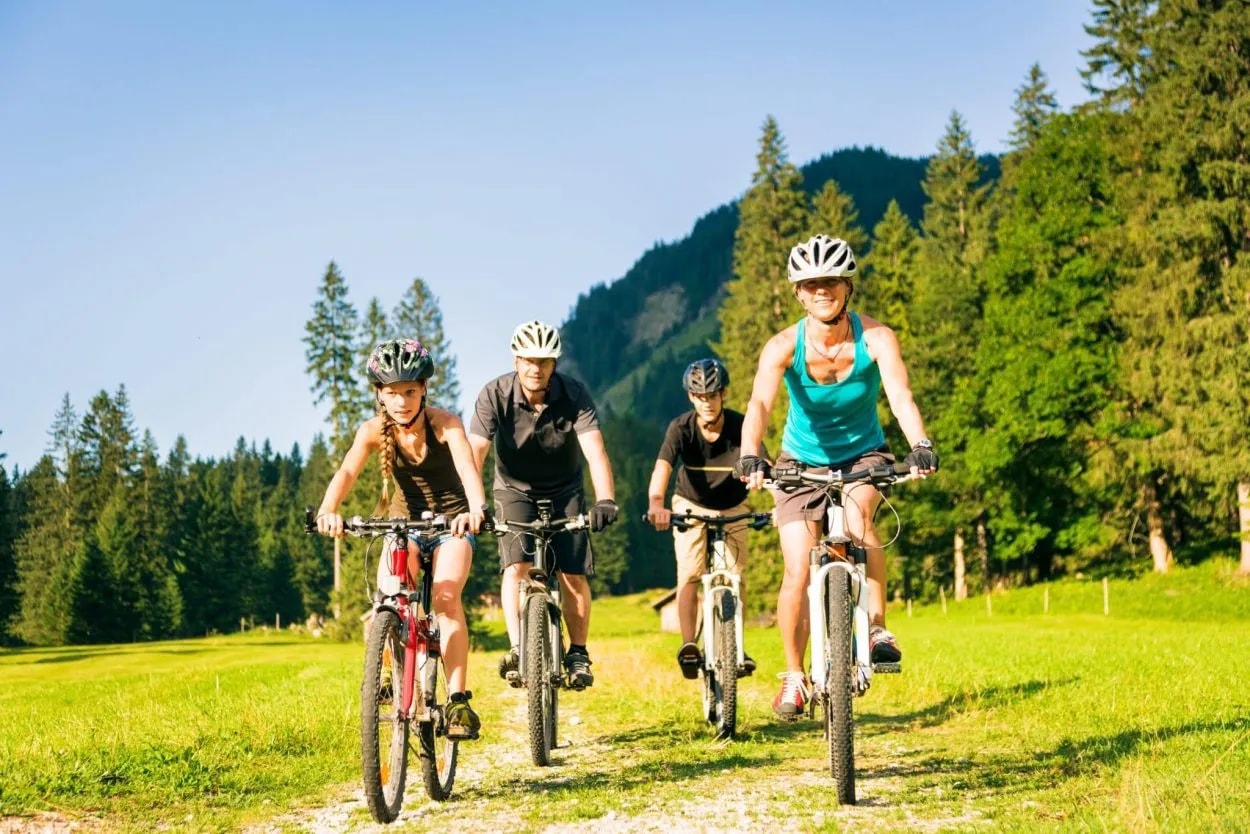 Family cycling on dirt path through grassy meadow with forested mountains in background