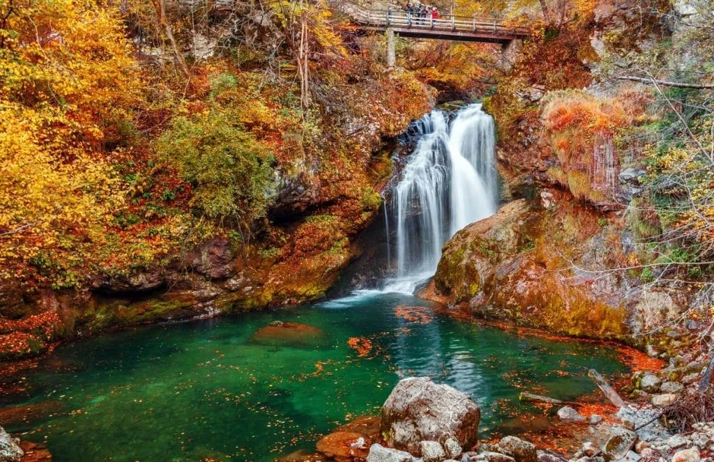 Vintgar Waterfall cascading into emerald pool surrounded by autumn foliage and wooden footbridge.