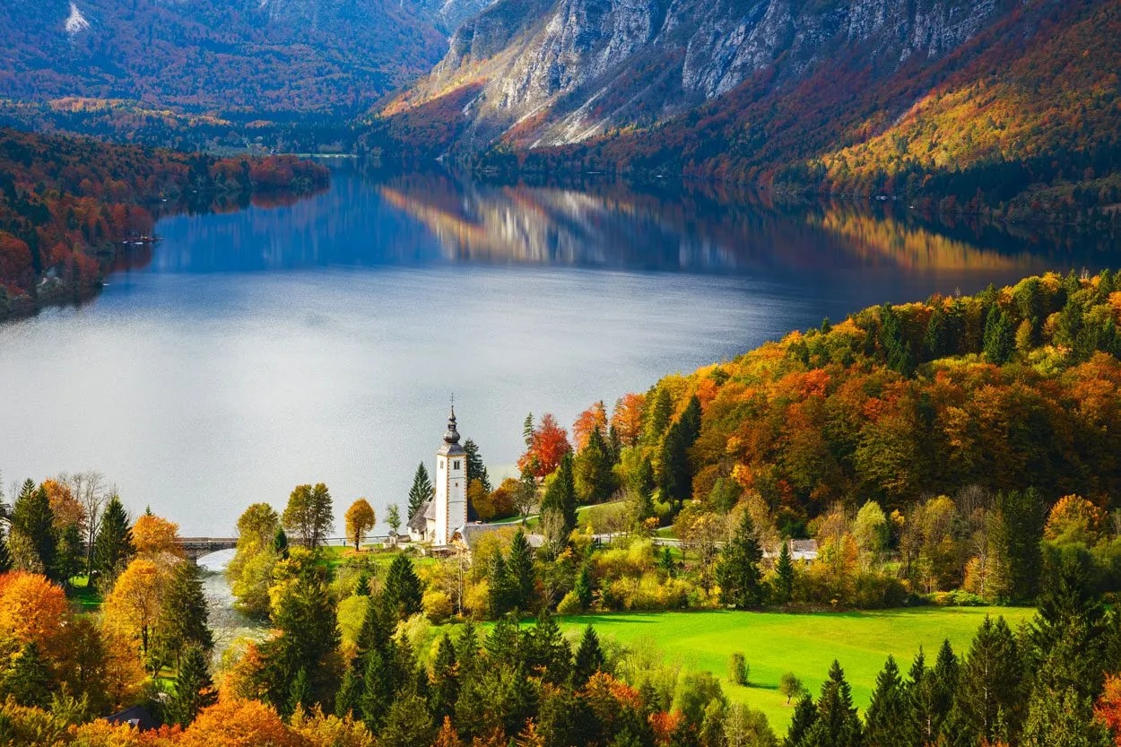 Panoramic view of Bohinj Lake with church and mountains in autumn foliage.