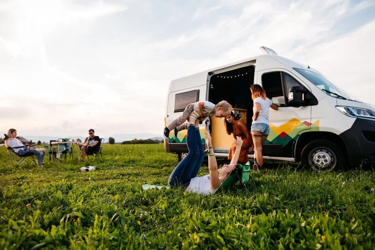 Family camping with camper van in grassy field; woman lifts child overhead.