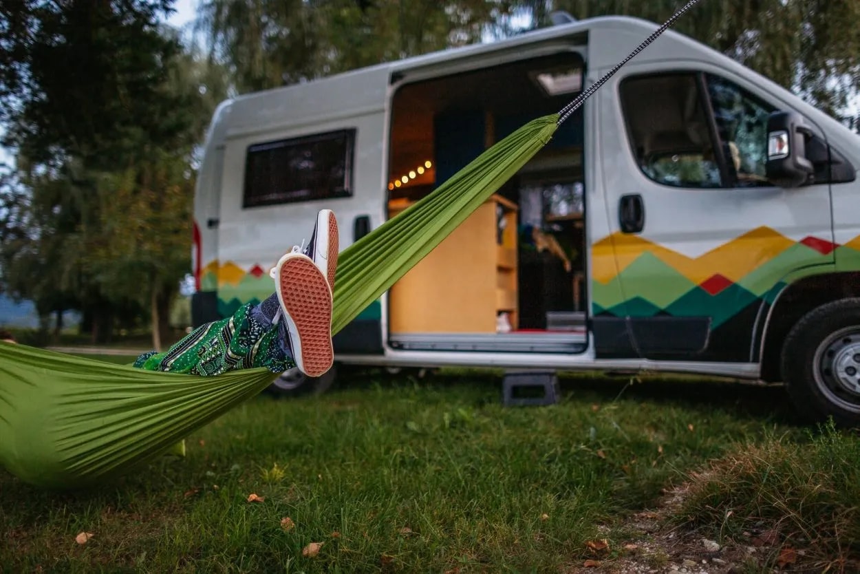 Person relaxing in green hammock next to camper van with mountain graphics parked on grass.