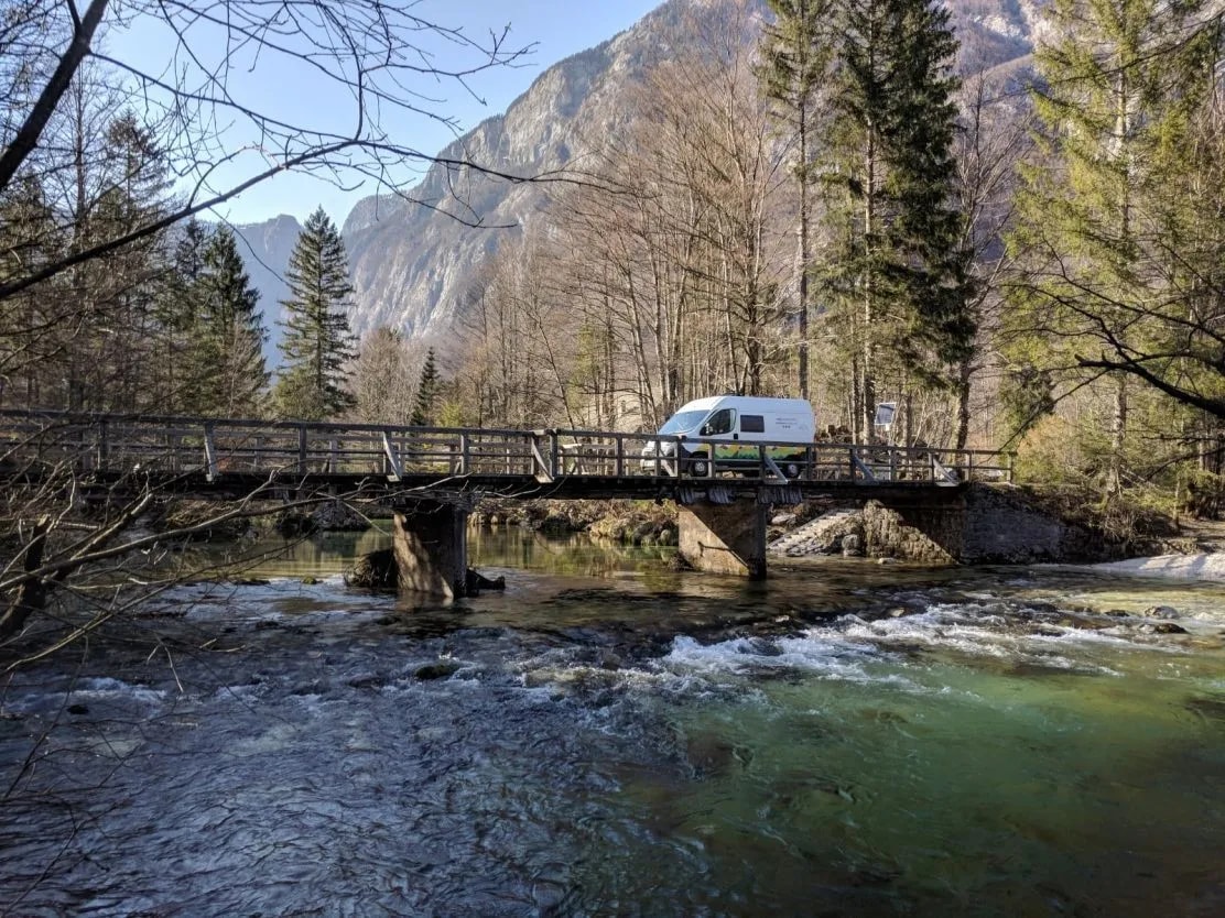 Camper van crossing wooden bridge over rushing river with mountains and forest background