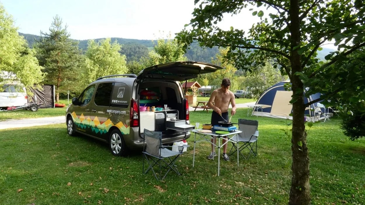 Man cooking outdoors at campsite next to camper van with open trunk in grassy, wooded area.