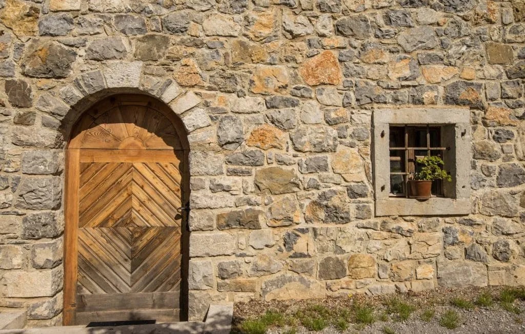 Stone house wall with arched wooden door and small window featuring a potted plant