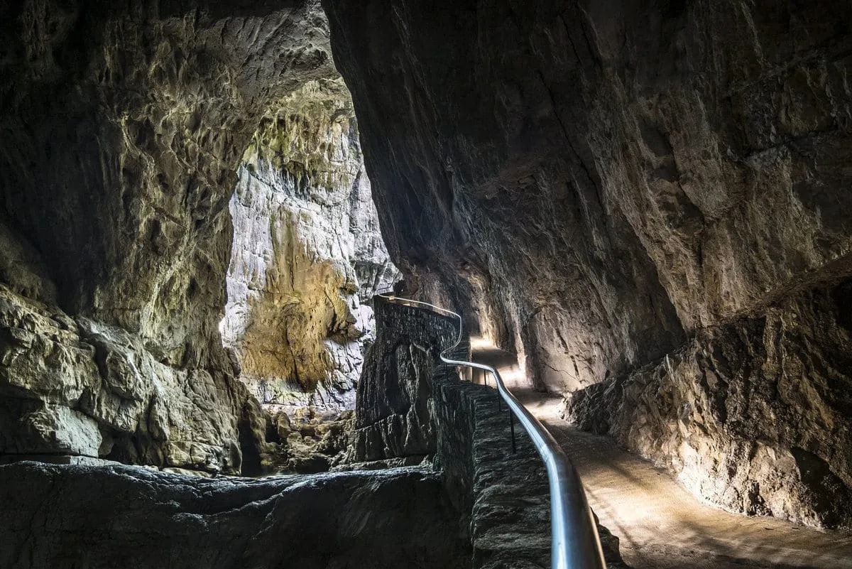 Winding stone pathway with metal railing inside the illuminated Skocjan Caves cavern.