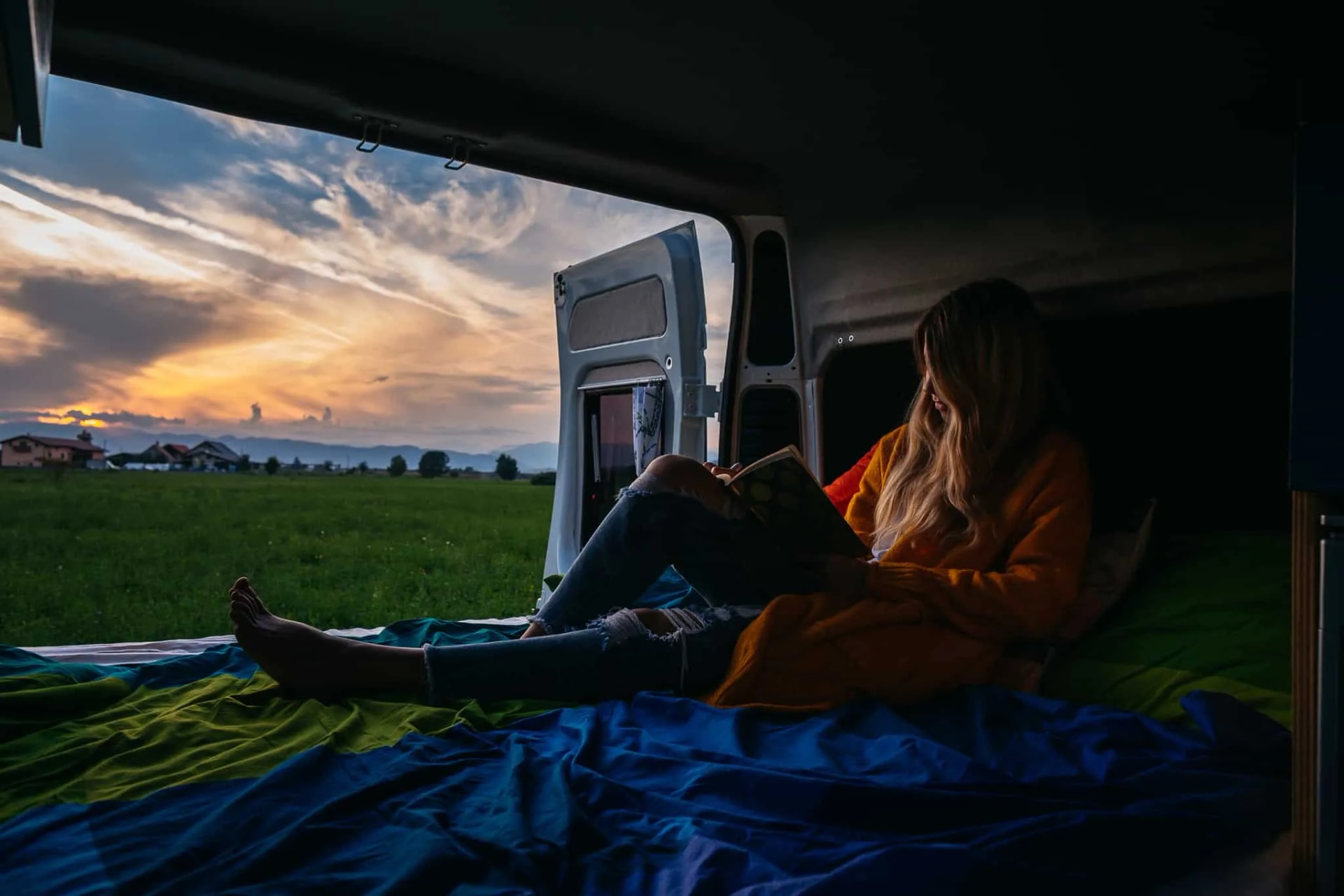 Woman reading inside camper van looking out at sunset over green field and distant houses.
