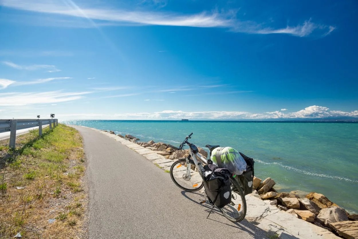 Bicycle touring along paved path next to turquoise sea under bright blue sky, Slovenian coast.
