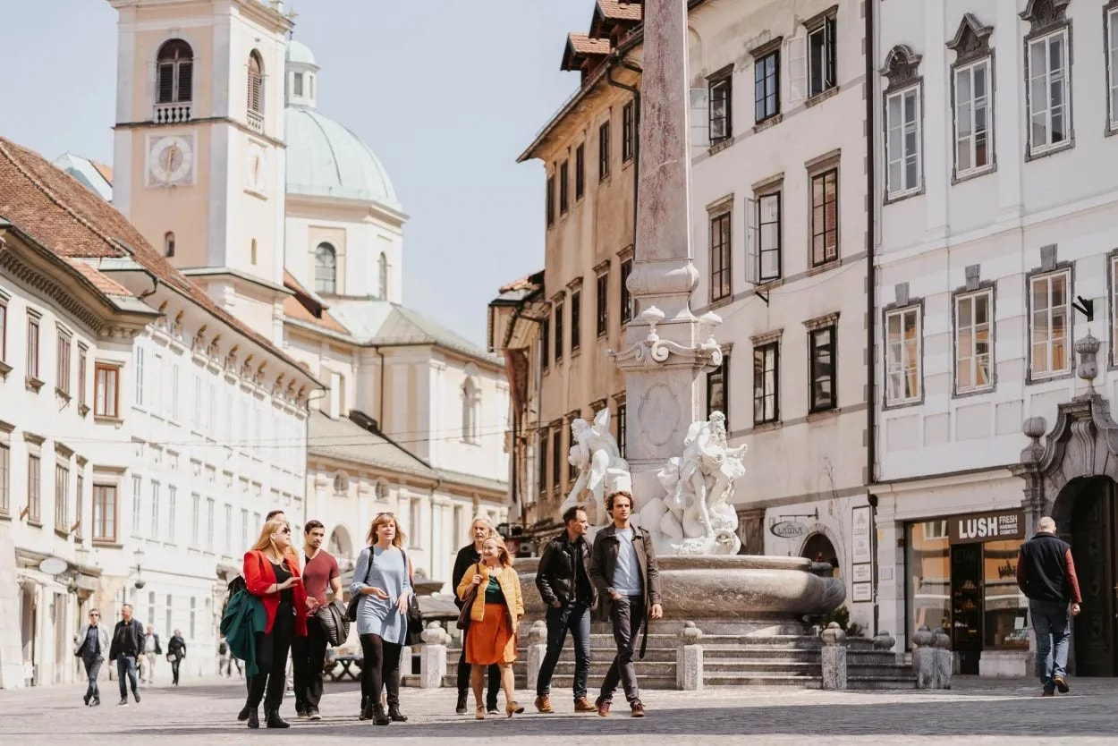 Tourists walking in Ljubljana old town square near fountain and church dome.
