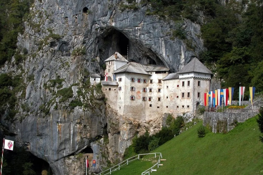 Predjama Castle built into a massive cliff face with green hillside and colorful flags.