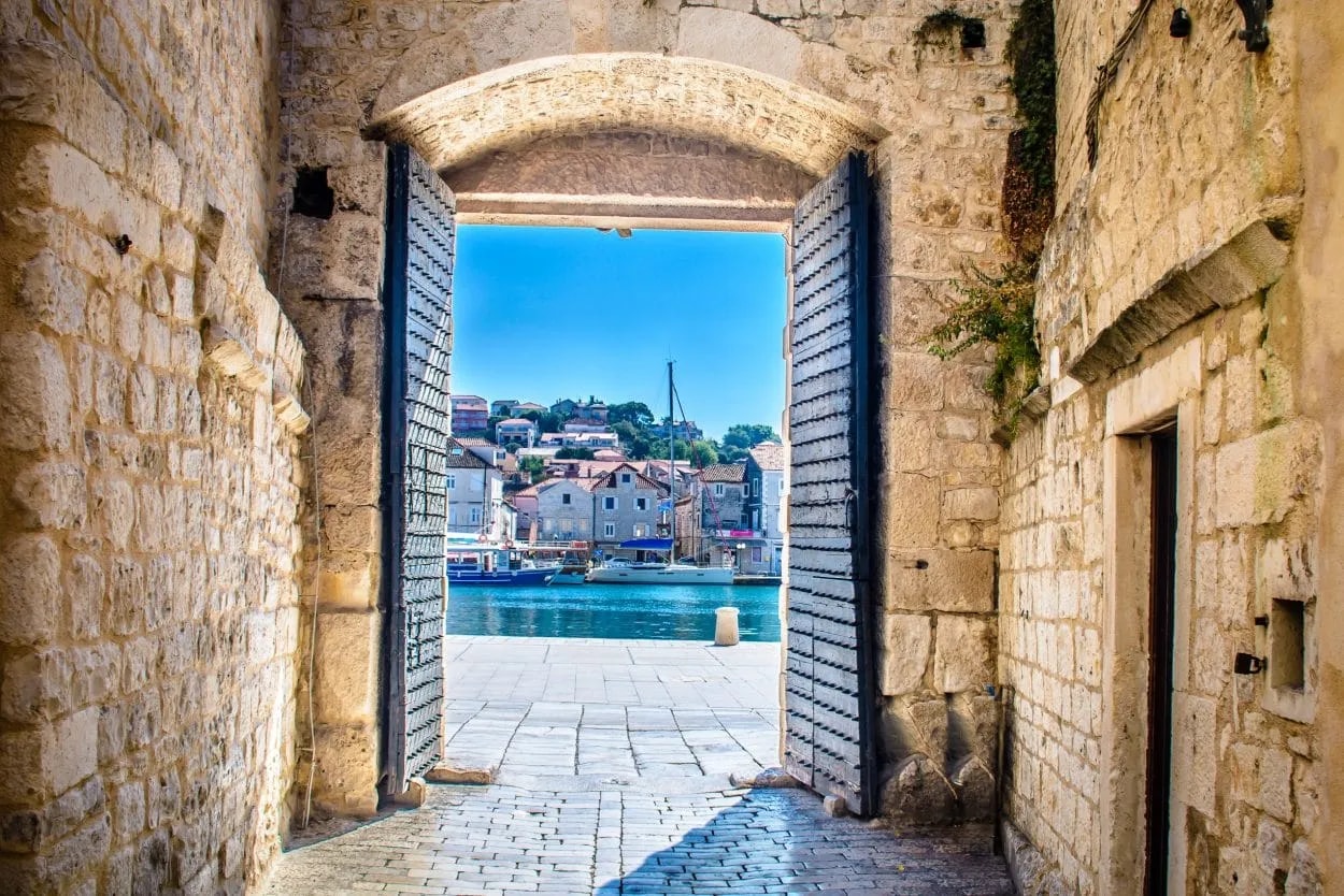 Stone archway with studded gates opening to a sunny harbor view in Trogir, Croatia.