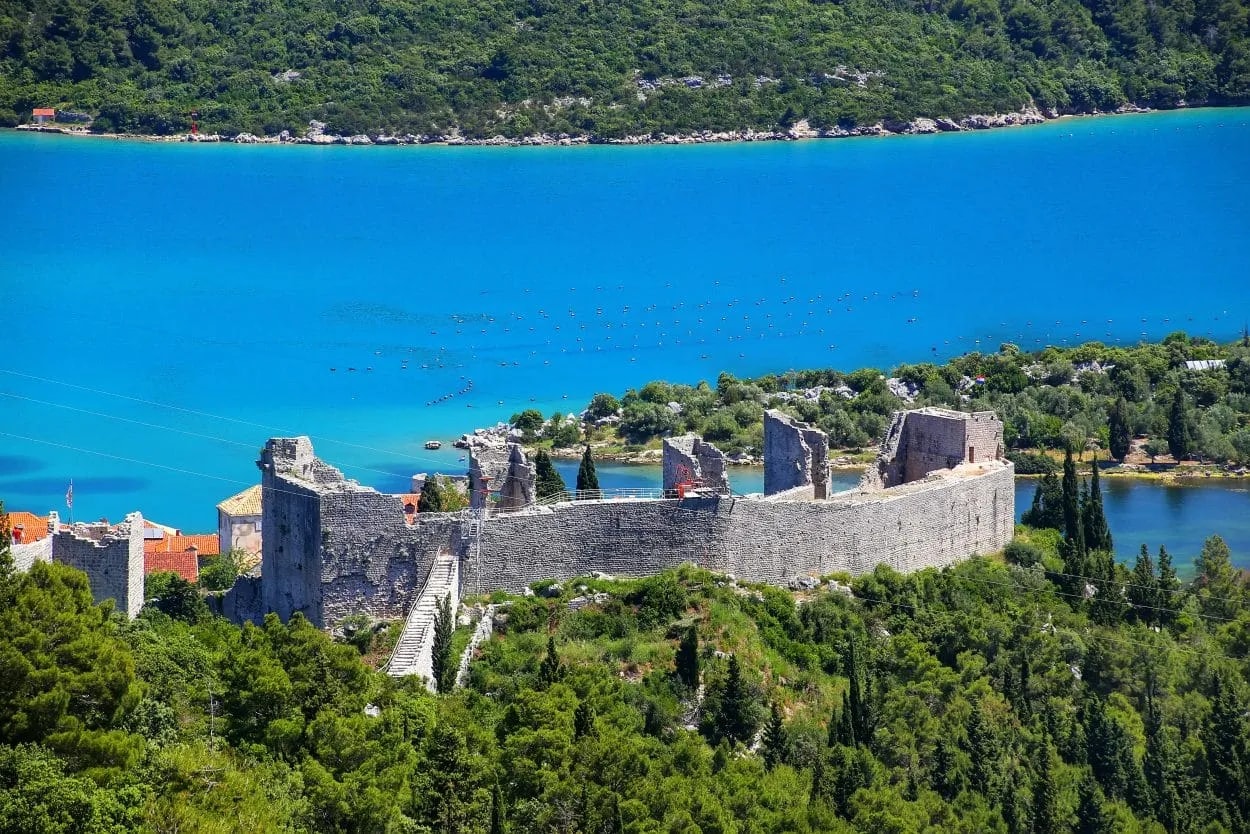 Stone fortification ruins overlooking bright blue coastal water surrounded by lush green forest.