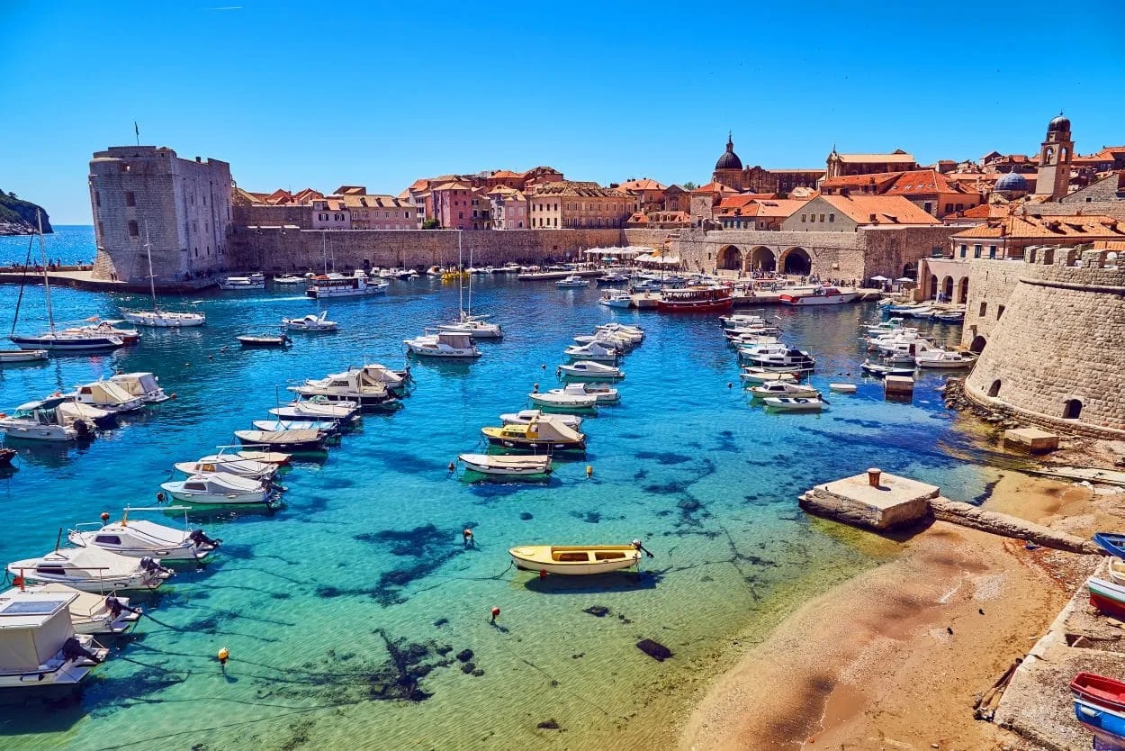 Boats moored in clear turquoise water outside the historic stone walls of Dubrovnik harbor.