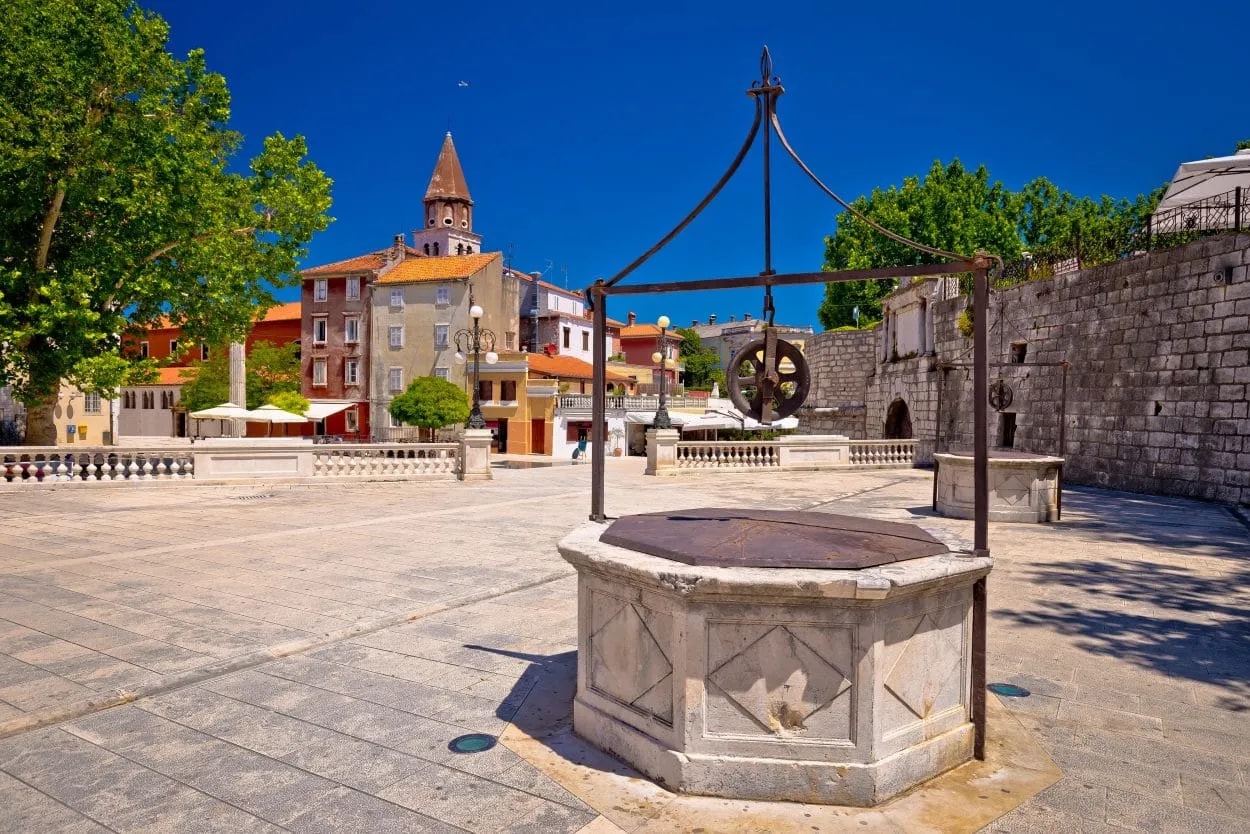 Historic stone well in a paved square with colorful buildings and a church spire in Zadar.
