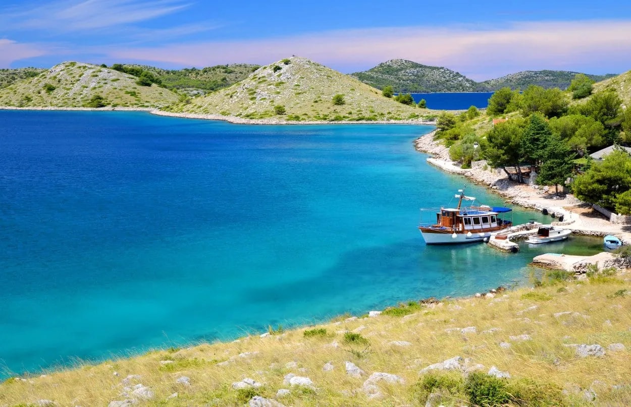 Boat docked in turquoise cove near rocky, scrub-covered hills of Kornati Islands.