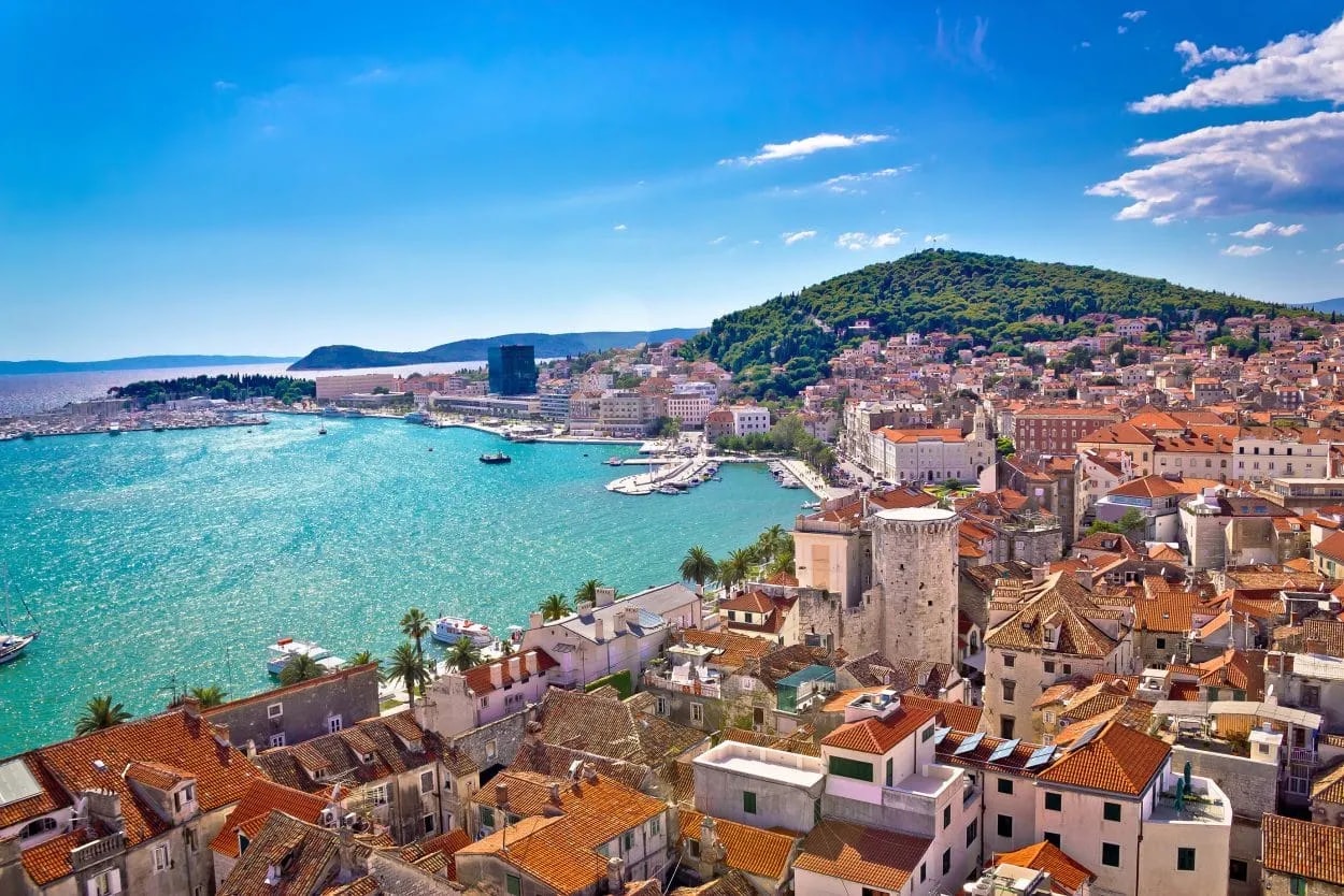 Coastal city with terracotta roofs and a harbor, backed by a lush green hill under a bright blue sky.