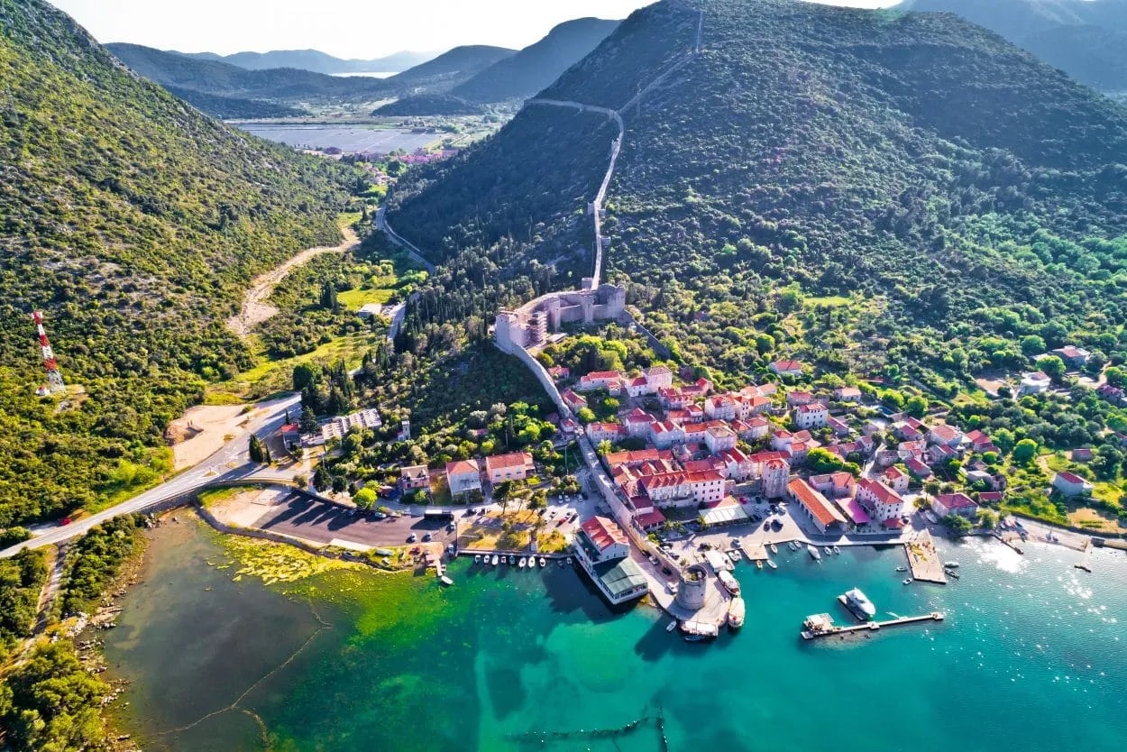 Coastal town with red-roofed buildings, harbor, and long defensive wall up green mountains.