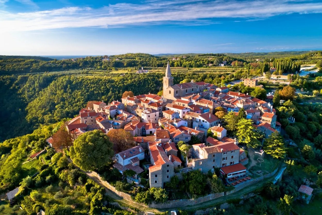 Hilltop medieval town of Grožnjan with terracotta roofs surrounded by lush green landscape under a blue sky.