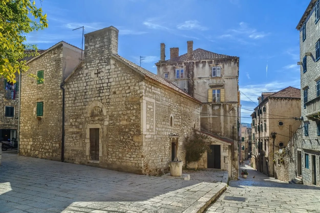 Stone church and historic buildings lining a steep cobblestone street in Šibenik under a bright blue sky.